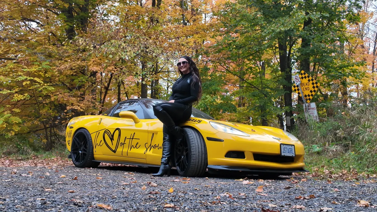 Chelsea Walsh seated on the front of her yellow Corvette C6, surrounded by autumn colour.