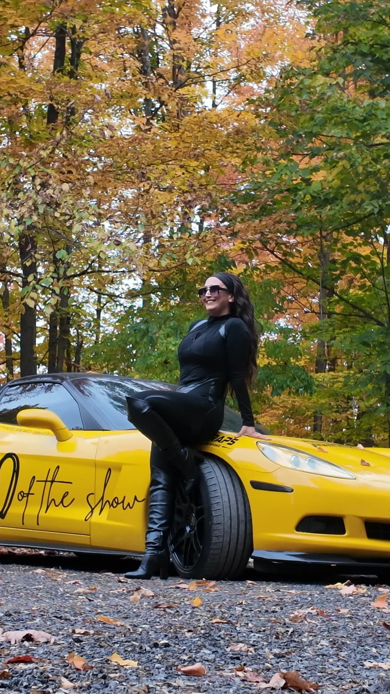 Chelsea Walsh smiling, seated on the fender of her yellow Corvette C6.