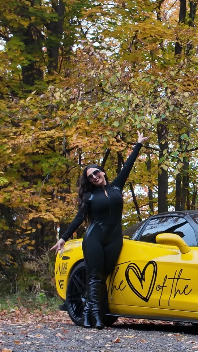 Chelsea waving beside the Corvette, autumn trees behind.
